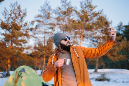 Téléchargez les photos : Après l'ascension, le touriste admire le coucher de soleil tout en se tenant près de la tente avec une tasse de thé ou de café et prend une photo sur son téléphone. - en image libre de droit