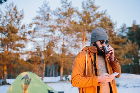 Téléchargez les photos : Après l'ascension, le touriste admire le coucher de soleil tout en se tenant près de la tente avec une tasse de thé ou de café et prend une photo sur son téléphone. - en image libre de droit