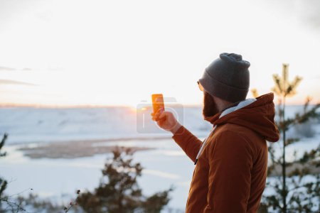 Photo for A hiker enjoys the sunset in the winter mountains and takes a photo on his phone. - Royalty Free Image