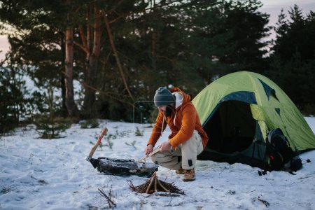 Téléchargez les photos : Un homme assis près de sa tente prépare de la laine de bois avec un couteau pour allumer un feu dans une forêt d'hiver au coucher du soleil. Concept de survie hivernale. - en image libre de droit