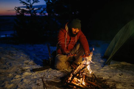 Foto de Un hombre se sienta cerca de una fogata, calentándose junto al fuego en un bosque de invierno al atardecer. - Imagen libre de derechos