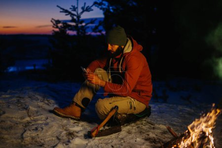 Téléchargez les photos : Un randonneur vérifie son téléphone tout en se réchauffant par un feu de camp la nuit dans les montagnes. - en image libre de droit