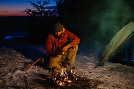 Téléchargez les photos : Image d'un jeune homme randonneur cuisinant un dîner au coin d'un feu de camp dans les montagnes en hiver. Randonneur masculin avec une barbe assise près de la tente après le coucher du soleil, faisant un feu de camp après la randonnée. Voyage, mode de vie concept. - en image libre de droit