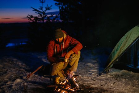 Photo for Image of hiker young man cooking dinner by a campfire in the mountains in winter. Male hiker with a beard sitting by the tent after sunset, making a campfire after hiking. Travel, lifestyle concept. - Royalty Free Image