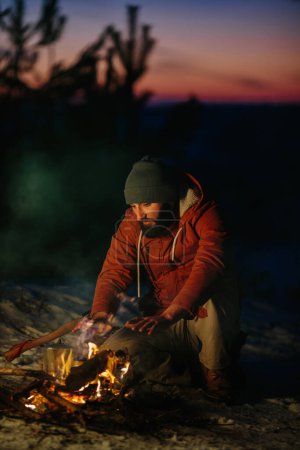 Téléchargez les photos : Un homme est assis près d'un feu de camp, se réchauffant par le feu dans une forêt d'hiver au coucher du soleil. - en image libre de droit