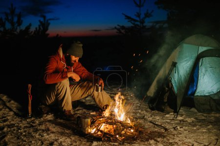 Téléchargez les photos : Un jeune homme barbu se repose près du feu de camp de nuit dans la forêt d'hiver. Le concept de survie et de randonnée dans la nature. - en image libre de droit