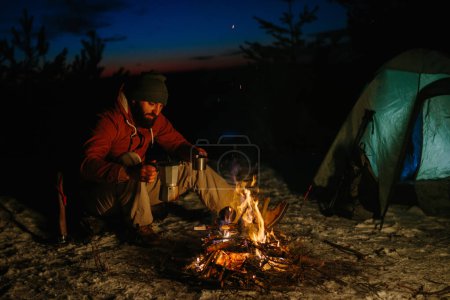 Foto de Un joven barbudo hace un delicioso café en una hoguera mientras disfruta de la puesta de sol en un bosque de invierno. El concepto de supervivencia y senderismo en la naturaleza. - Imagen libre de derechos