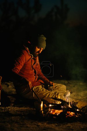 Foto de Un joven barbudo descansa junto a la fogata nocturna en el bosque invernal. El concepto de supervivencia y senderismo en la naturaleza. - Imagen libre de derechos