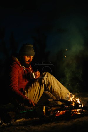 Foto de Un joven barbudo descansa junto a la fogata nocturna en el bosque invernal. El concepto de supervivencia y senderismo en la naturaleza. - Imagen libre de derechos