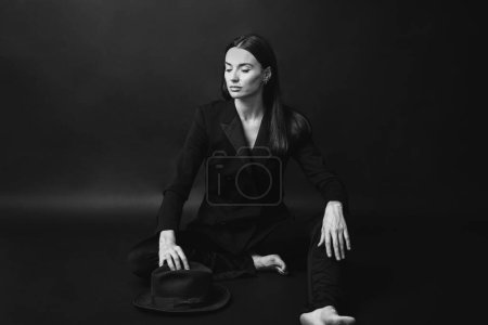Téléchargez les photos : Portrait d'une femme moderne et élégante. Une brune en costume et chapeau pose pieds nus sur un fond noir. - en image libre de droit