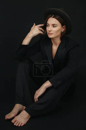 Photo for Portrait of a modern stylish woman. A brunette in a suit and hat poses barefoot on a black background. - Royalty Free Image
