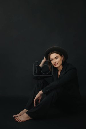 Photo for Portrait of a modern stylish woman. A brunette in a suit and hat poses barefoot on a black background. - Royalty Free Image