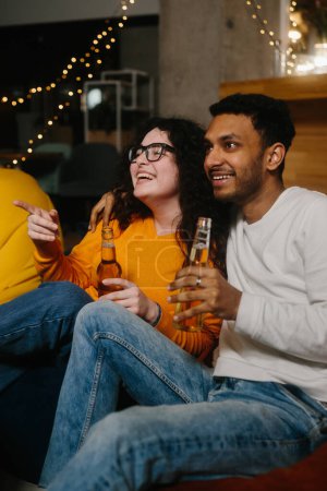 Foto de Un par de amigos multirraciales están viendo una película de comedia mientras están sentados en bolsas de frijoles blandos mientras beben cerveza. - Imagen libre de derechos