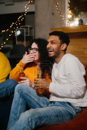 Foto de Un par de amigos multirraciales están viendo una película de comedia mientras están sentados en bolsas de frijoles blandos mientras beben cerveza. - Imagen libre de derechos