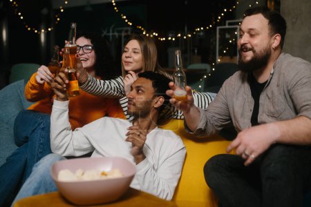 Foto de Un grupo de amigos come bocadillos y bebe cerveza mientras ven la televisión. - Imagen libre de derechos
