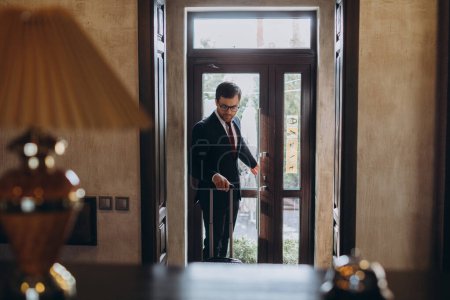 Photo for Pleasant smiling man in classic suit passing the entrance of a busines hotel dragging his baggage - Royalty Free Image