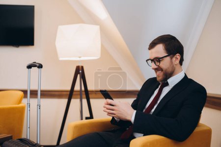 Photo for A businessman checks his phone while sitting in a comfortable chair in a hotel room. - Royalty Free Image