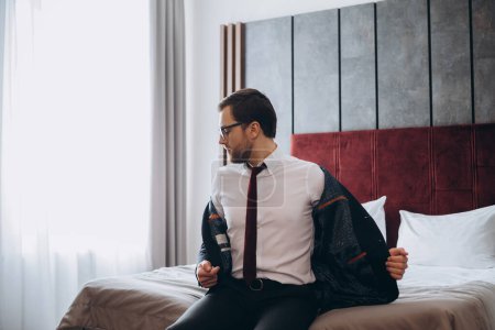 Business man with suitcase taking off jacket while sitting on bed in hotel room