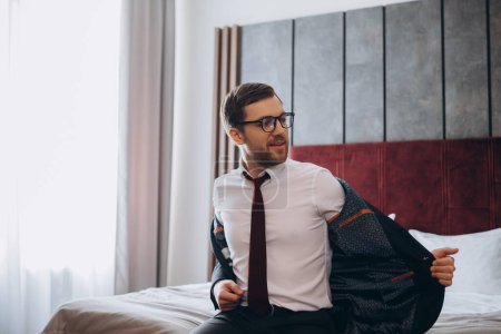 Geschäftsmann mit Koffer zieht Jacke aus, während er auf Bett im Hotelzimmer sitzt
