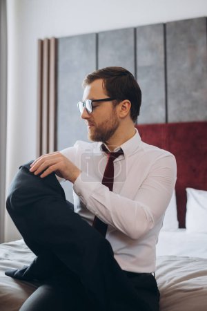 Téléchargez les photos : Homme d'affaires avec valise enlevant la veste assis sur le lit dans la chambre d'hôtel - en image libre de droit