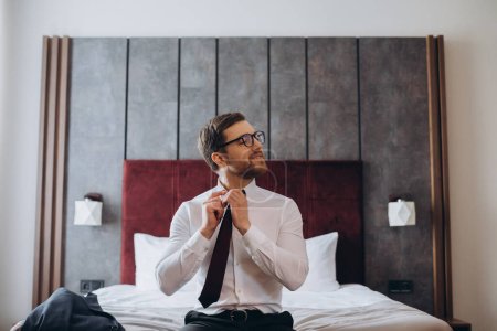 Téléchargez les photos : Jeune homme d'affaires enlevant sa cravate assis sur un lit dans une chambre d'hôtel. - en image libre de droit