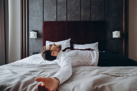 Téléchargez les photos : Homme d'affaires épuisé se reposant sur le lit après un long vol aérien, décalage horaire. Homme en costume d'affaires couché avec les bras tendus sur le matelas dans la chambre d'hôtel. - en image libre de droit