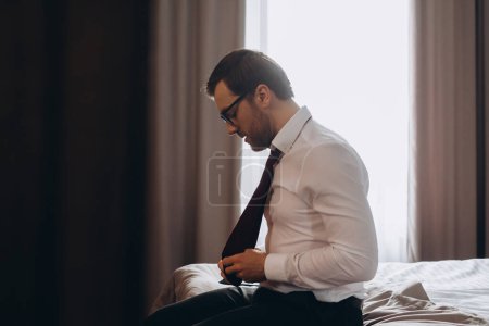 Junger Geschäftsmann zieht seine Krawatte aus, während er in einem Hotelzimmer auf einem Bett sitzt.