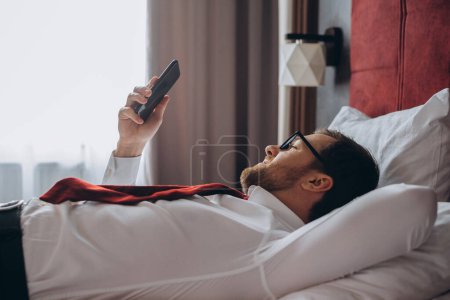 Téléchargez les photos : Un homme d'affaires repose allongé sur un lit dans une chambre d'hôtel. Un jeune homme utilise son smartphone tout en se reposant. - en image libre de droit