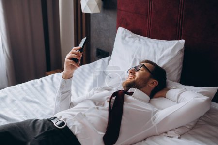 Photo for A businessman rests lying on a bed in a hotel room. A young man uses his smartphone while resting. - Royalty Free Image