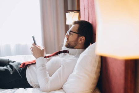Téléchargez les photos : Jeune homme d'affaires travaillant dans une chambre d'hôtel avec son téléphone portable - en image libre de droit