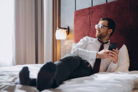 Foto de Hombre de negocios alegre usando teléfono sentado en la habitación de hotel en tri de negocios - Imagen libre de derechos