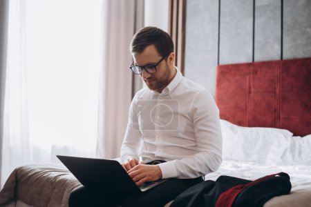 Photo for Young businessman working with computer sit on bed in hotel room - Royalty Free Image