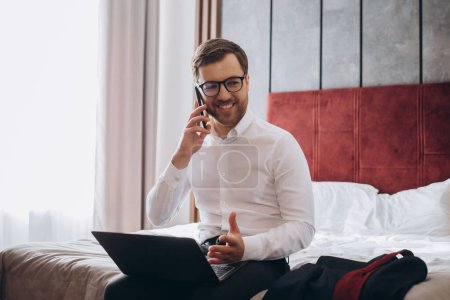 Téléchargez les photos : Beau homme d'affaires faisant téléphone tout en étant assis sur le lit de la chambre d'hôtel et en utilisant un ordinateur portable pendant le voyage d'affaires - en image libre de droit