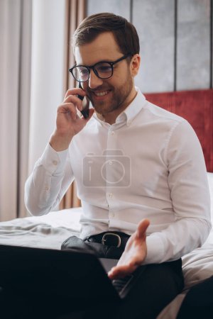 Beau homme d'affaires faisant téléphone tout en étant assis sur le lit de la chambre d'hôtel et en utilisant un ordinateur portable pendant le voyage d'affaires