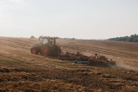 Téléchargez les photos : Un tracteur moderne avec une herse à disque traînée lourde laboure un champ de blé au coucher du soleil. - en image libre de droit