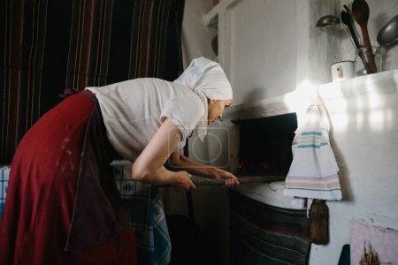 Photo for A woman in Ukrainian national dress works near a wood-burning stove. The traditional process of baking bread at home. - Royalty Free Image