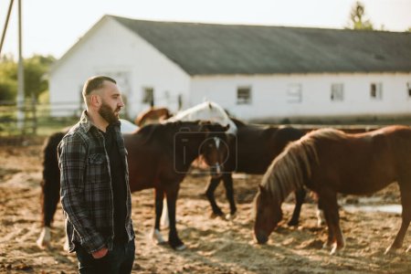 Téléchargez les photos : Portrait d'un jeune ouvrier agricole, sur fond de clôture à chevaux. Un jeune fermier s'occupe des chevaux dans les écuries. - en image libre de droit