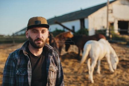 Foto de Un joven granjero cerca de una manada de caballos en una granja. Retrato de un joven barbudo sobre el fondo de los caballos al atardecer. - Imagen libre de derechos
