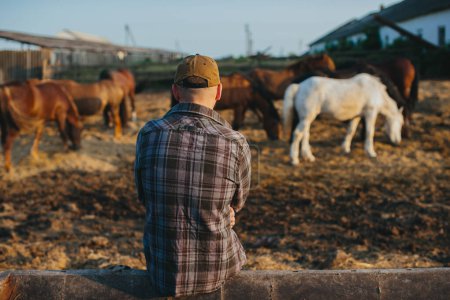 Photo for Portrait of a young farm worker, on the background of a fence with horses. A young farmer takes care of horses in the stables. - Royalty Free Image