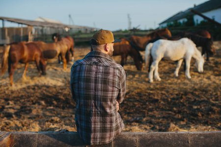 Foto de Retrato de un joven campesino, sobre el fondo de una cerca con caballos. Un joven agricultor se encarga de los caballos en los establos. - Imagen libre de derechos