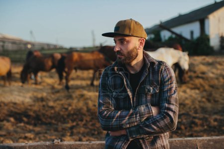 Téléchargez les photos : Portrait d'un jeune ouvrier agricole, sur fond de clôture à chevaux. Un jeune fermier s'occupe des chevaux dans les écuries. - en image libre de droit