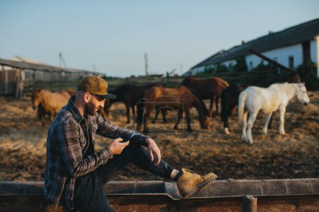 Téléchargez les photos : Un jeune homme avec un téléphone dans les mains sur le fond d'un enclos avec des chevaux. Un homme se repose après une journée de travail à la ferme, coucher de soleil. - en image libre de droit