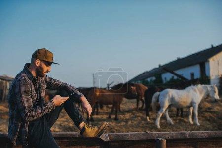 Foto de Un joven con un teléfono en sus manos en el fondo de un paddock con caballos. Un hombre descansa después de un día de trabajo en la granja, puesta de sol. - Imagen libre de derechos