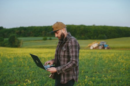 Photo for Caucasian young male farmer in hat standing in field and typing on keyboard of laptop computer. Attractive man using device in margin in summer and browsing online. - Royalty Free Image