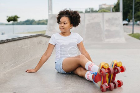 Téléchargez les photos : Mignon afro américaine fille roller skater assis dans le parc de la ville et souriant. - en image libre de droit