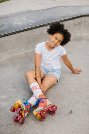 Téléchargez les photos : Mignon afro américaine fille roller skater assis dans le parc de la ville et souriant. - en image libre de droit