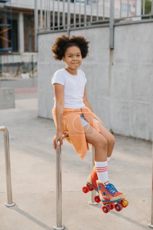 Téléchargez les photos : Jeune fille afro-américaine en patins à roulettes s'amusant à l'extérieur. La fille sourit en regardant la caméra. - en image libre de droit