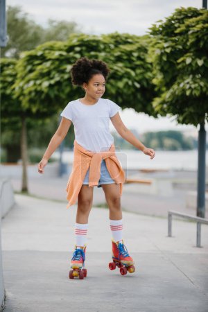 Téléchargez les photos : Afro-Américaine jeune fille patinage à roulettes en plein air, enfant jouant sur des patins à roulettes. - en image libre de droit