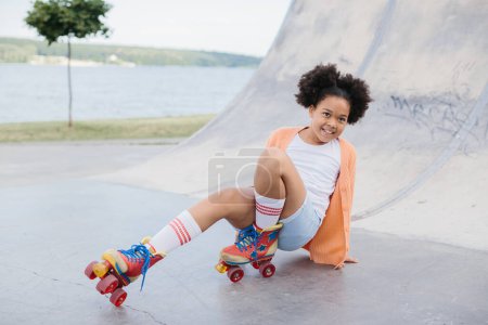 Foto de Linda chica afroamericana patinadora sentada en el parque de la ciudad y sonriendo. - Imagen libre de derechos