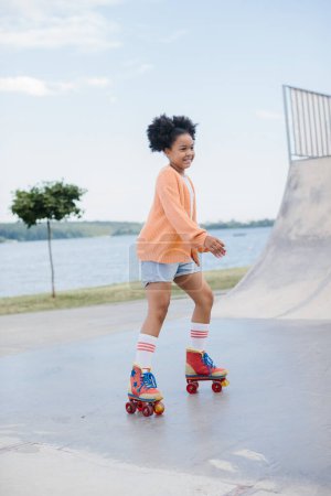 Téléchargez les photos : Petite fille afro-américaine mignonne chevauchant sur patins à roulettes dans un parc d'été. Enfant jouissant de sport en plein air dans le parc, drôle petite fille jouant sur le patin à roulettes sur la route. sport apprentissage concept outdoor - en image libre de droit